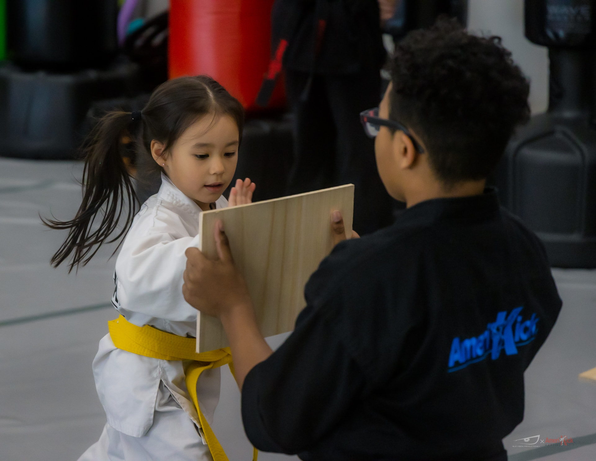 A young student in a white gi with a yellow belt striking a wooden board held by an instructor